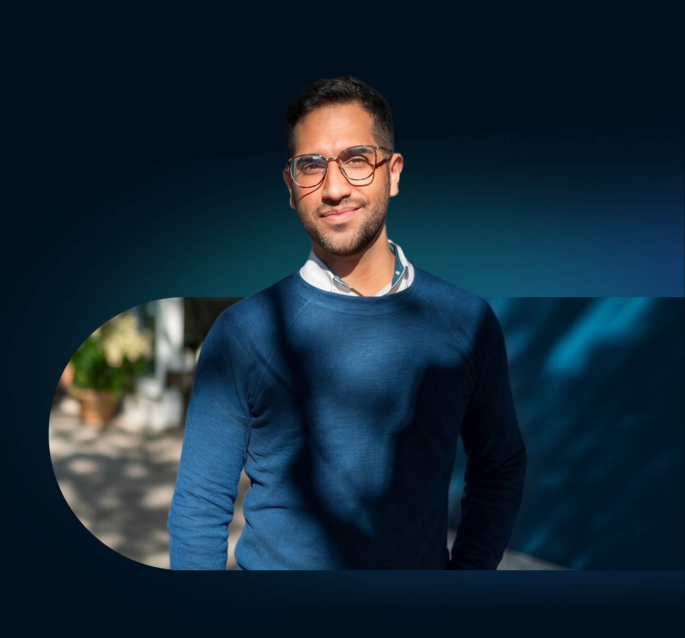 Portrait of a smiling man in glasses and a blue sweater, set against a dark blue background with a blurred outdoor scene layered behind him.